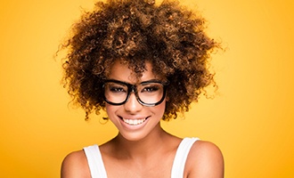 A young female with black glasses and a white tank top shows off her complete smile thanks to a single tooth dental implant
