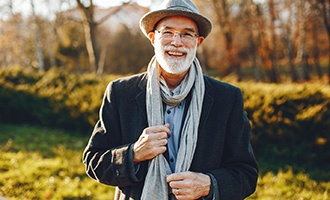 An older man wearing a coat, scarf, and hat smiles after a successful appointment with his implant dentist