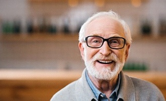 An older man with glasses smiles after receiving his implant dentures in Massapequa